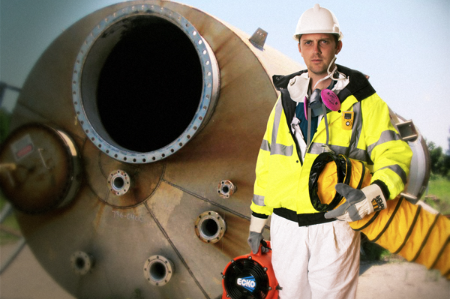 Worker standing in front on confined space entry point holding ventilation and fan, while wearing hard hat and other PPE.