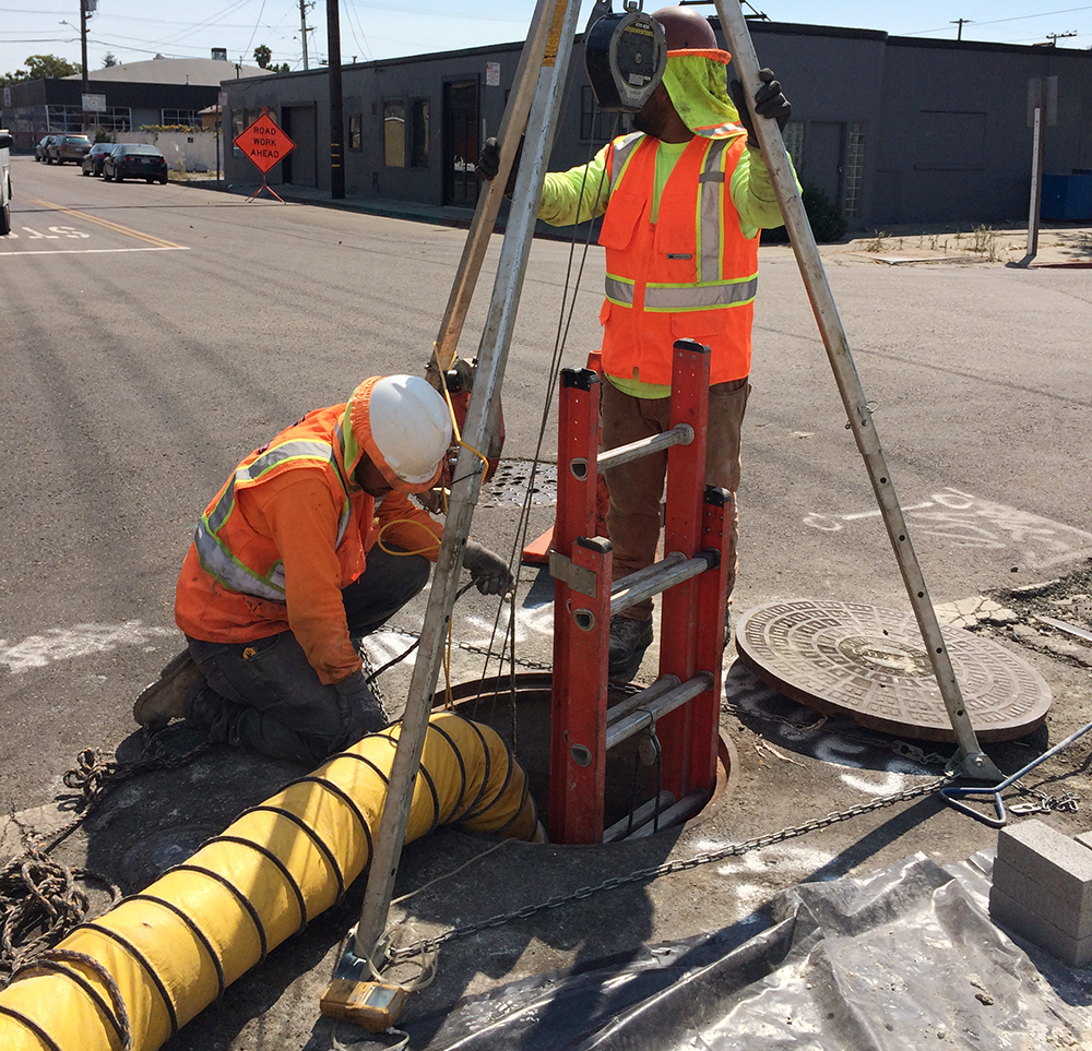 Workers using a tripod and ventilation over a manhole in the middle of a street.