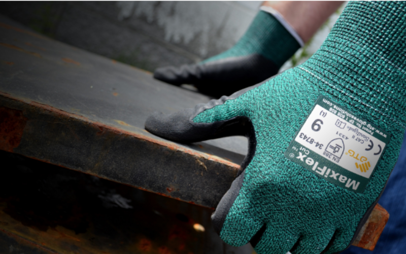 A person wearing green Maxiflex gloves grips a rusty metal surface. The gloves, highlighted in the foreground, indicate protection and industrial work.