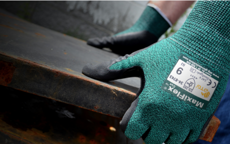 A person wearing green Maxiflex gloves grips a rusty metal surface. The gloves, highlighted in the foreground, indicate protection and industrial work.