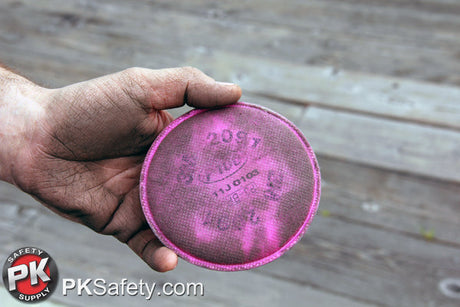 A dirty hand holds a pink 3M P100 respirator filter. The filter is also dirty, indicating thorough use.