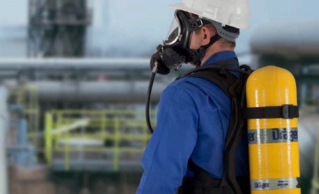 Worker wearing SCBA with yellow tank, helmet and full-face respirator looking towards an industrial plant.
