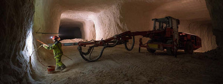 A miner in protective gear examines a tunnel wall, illuminated by soft light. A large drilling machine is parked nearby in the expansive underground space.