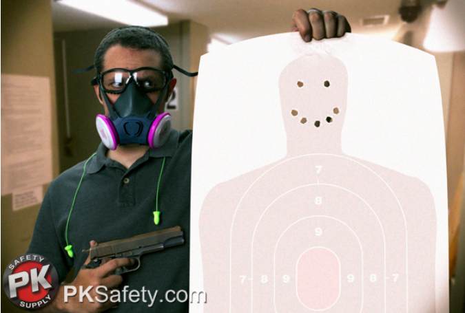 Man at gun range holding shot target and gun. Wearing respirator and safety glasses.