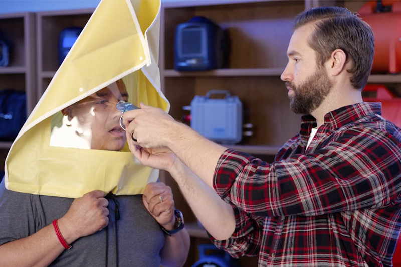 A man in a plaid shirt helps another person wearing a yellow emergency hood by adjusting the mask over their face. Shelves with emergency equipment are visible in the background.