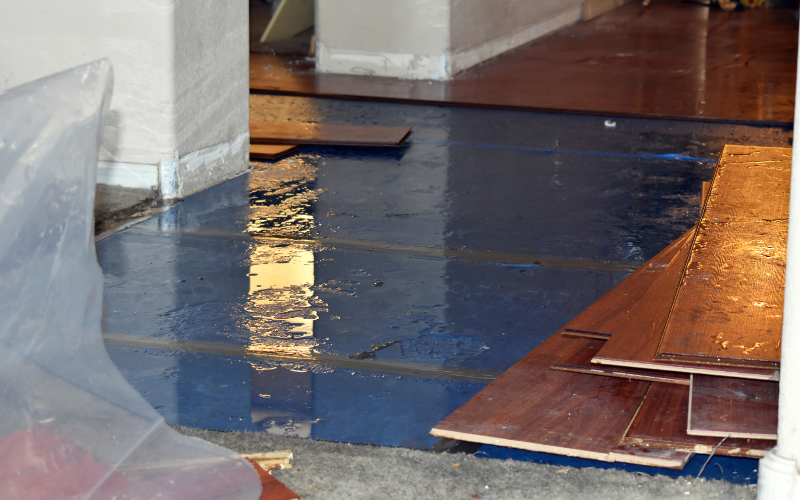 Flooded room with a wet blue floor, scattered wooden planks, and a transparent plastic sheet. The scene suggests an unfinished renovation or water damage.