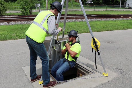Two workers wearing safety vests and helmets use a tripod system; one is being lowered into a manhole while the other operates the equipment beside a railway track.