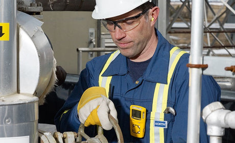 Worker wearing safety gear and gloves operates machinery outdoors. He is focused, uses a handheld device, and wears a hard hat, earplugs, and protective eyewear.