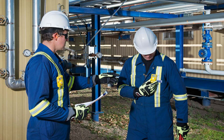 Two workers in blue coveralls and white hard hats discuss near industrial piping. One holds a clipboard, both appear focused and engaged in conversation.