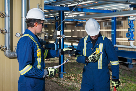 Two oil and gas workers with pipes behind them. One looks down at gas detector on his chest.