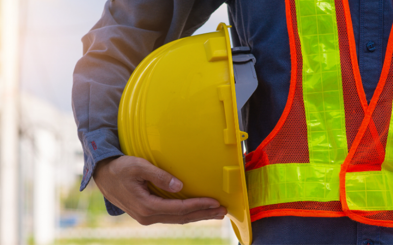 A person wearing a neon safety vest holds a yellow hard hat. The background is blurred, highlighting construction or worksite safety.