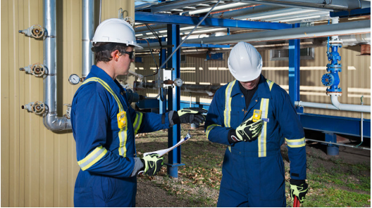 Two workers wearing blue coveralls and white hard hats with gas monitors clipped to their chests. One looks down at his.