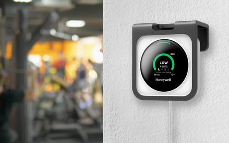 A digital air quality monitor displaying "LOW" in green is mounted on a white wall. Blurred gym equipment is visible in the background, indicating a fitness setting.