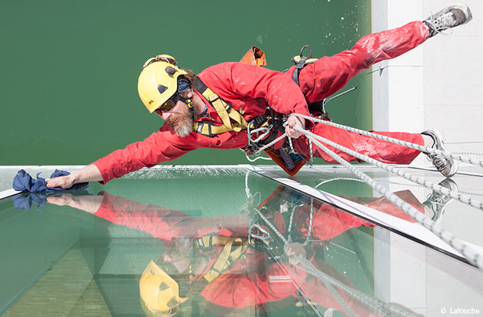 A window cleaner in a red suit and yellow helmet is suspended by ropes, cleaning a glass building surface. The scene conveys focus and skill.