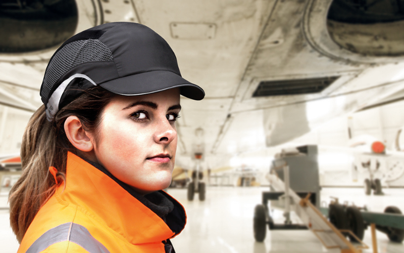 Young woman in an orange safety jacket and black bump cap stands confidently in an aircraft hangar, with a visible plane and maintenance equipment.