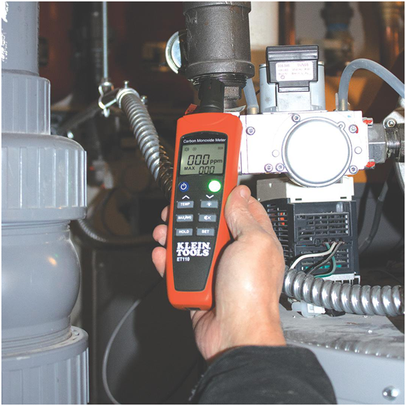 A hand holds a Klein Tools ET110 carbon monoxide meter by industrial pipes. The display shows "0.00 PPM," indicating safe levels. The setting appears mechanical and secure.