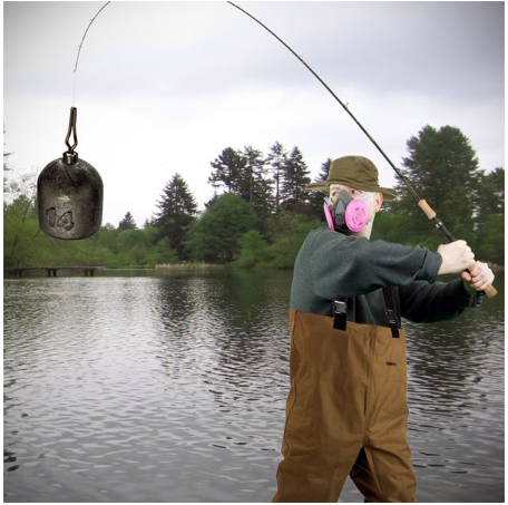 Man wearing fishing gear and half-mask respirator with a lead weight on his fishing line and a lake in the background.