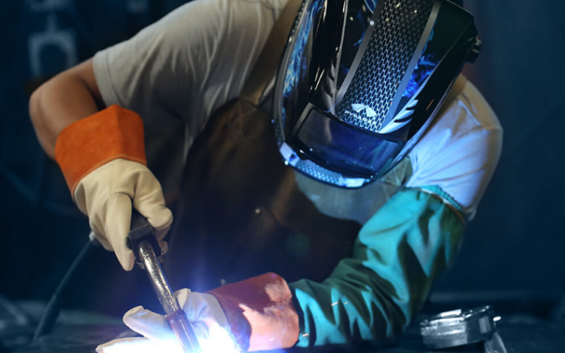 A welder in protective gear, including a helmet and gloves, works intently with a bright welding torch, casting a sharp blue glow.