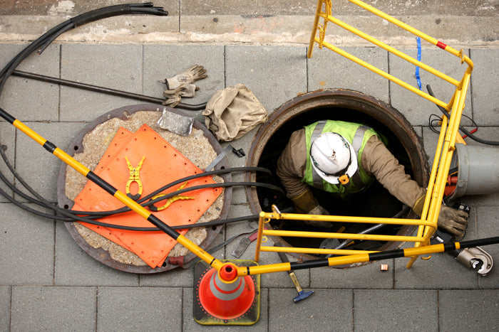 Worker in a safety vest and helmet descending into a manhole on a sidewalk. Surrounding safety barriers and orange cones indicate ongoing maintenance work.