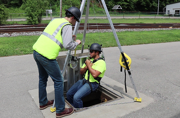 Two men wearing safety vests and helmets are engaged in work on a manhole, demonstrating teamwork in a construction setting. One man is being lowered into the manhole with a safety tripod and winch.