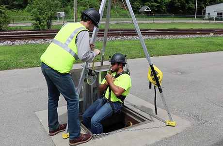 Two men wearing safety vests and helmets are engaged in work on a manhole, demonstrating teamwork in a construction setting. One man is being lowered into the manhole with a safety tripod and winch.