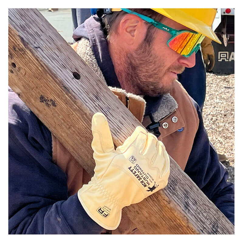 A construction worker in a yellow hard hat, reflective sunglasses, dark hoodie, and brown vest carries a wooden beam while wearing MCR Safety Mustang HiDex Kevlar Lined Leather Driver MU3664K gloves.