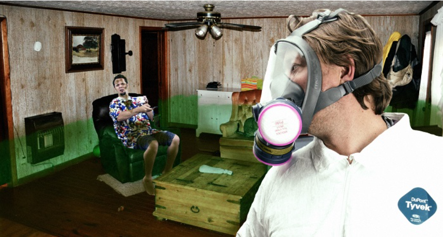Man wearing full-face respirator and hazmat suit looks towards a man sitting on a chair in a filthy room with a green gas floating around.
