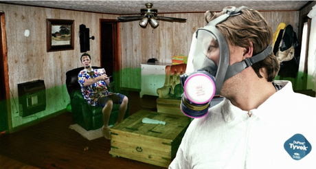 Man wearing full-face respirator and hazmat suit looks towards a man sitting on a chair in a filthy room with a green gas floating around.