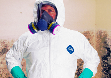 Man wearing hazmat suit and full-face respirator with moldy wall behind him.