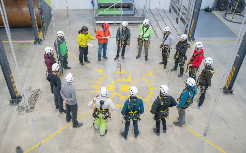 A group of people in safety gear and helmets stand in a circle on a marked floor in an industrial training facility, indicating teamwork and focus.