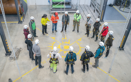 A group of people in safety gear and helmets stand in a circle on a marked floor in an industrial training facility, indicating teamwork and focus.