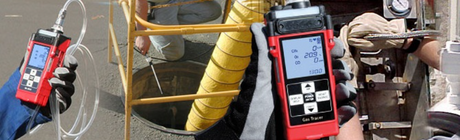 Photo of hands holding red gas monitors with manhole in the background.