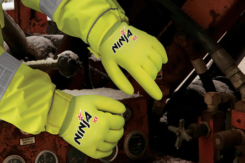 Two hands wearing bright yellow-green gloves labeled "Ninja Ice" are operating controls on a red industrial machine covered with patches of snow.