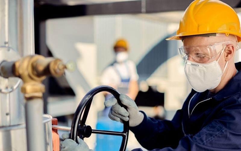 Worker in a hard hat and mask operates industrial equipment, turning a large valve. Background shows another worker, suggesting a factory setting.