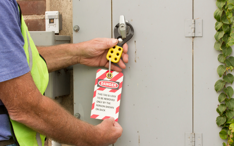 A person in a safety vest attaches a lock-out tag with a "Danger" sign to a gray electrical panel, emphasizing caution and security protocols.