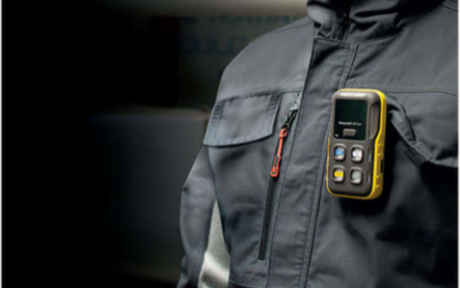 A person wearing a dark jacket with the yellow handheld BW Icon gas detection device clipped to the chest pocket. The device has a small screen and several buttons.