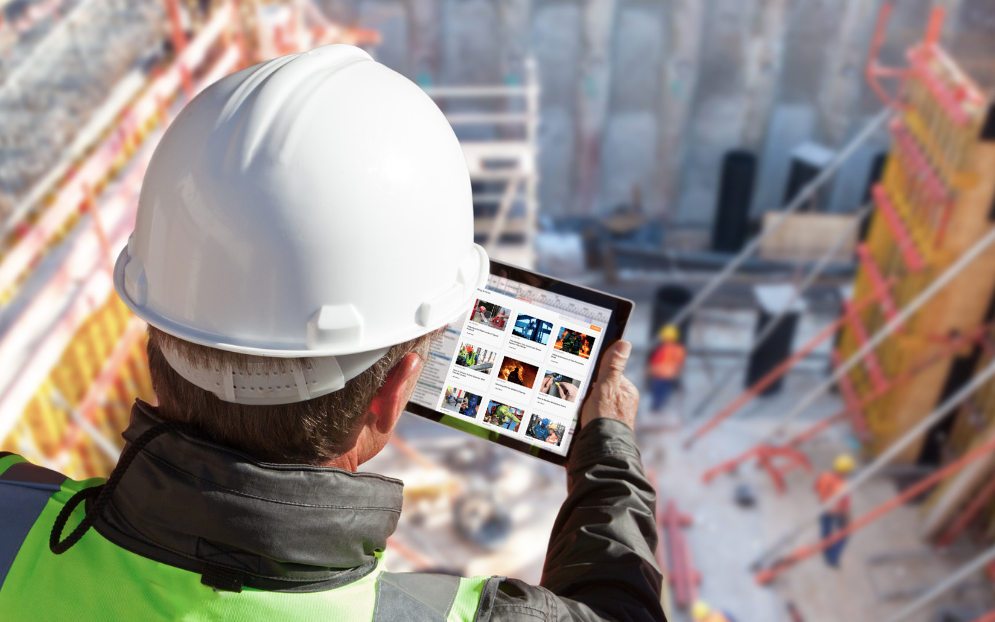 A construction worker in a hard hat and reflective vest uses a tablet at a building site. The scene is busy with scaffolding and construction materials.