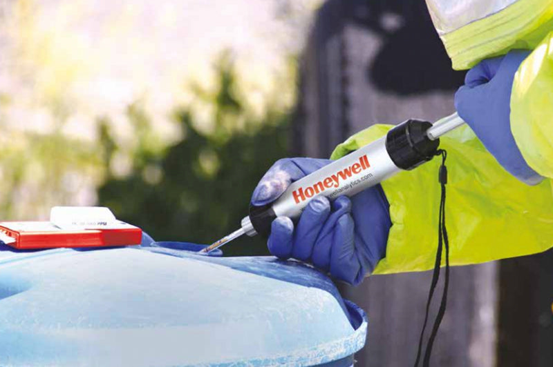 A person wearing a yellow protective suit and blue gloves uses a Honeywell-branded device to test or sample the contents of a large blue container outdoors.