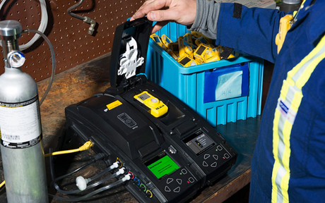 A person in a blue and yellow uniform calibrates a gas detector on a table with a gas cylinder. A blue bin with equipment is nearby.