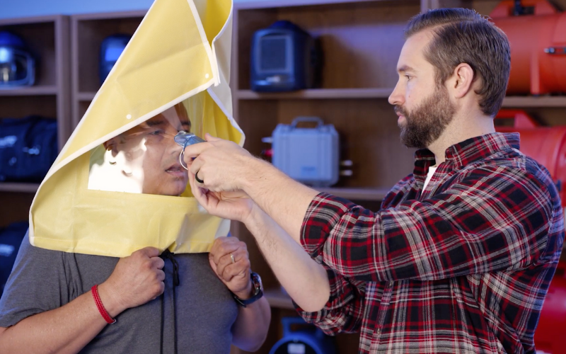 A man in a plaid shirt assists another man wearing a yellow emergency hood with a transparent face panel. Background shows shelves with emergency equipment.