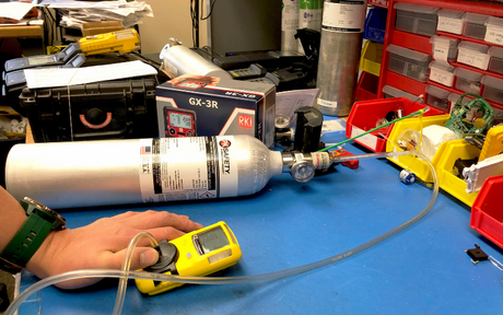 A worker tests a yellow gas detector using a calibration gas cylinder on a blue workbench. Tools and equipment are organized in the background.