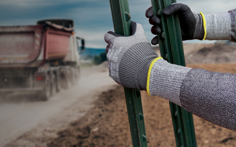 Person wearing protective gloves grips green metal posts on a dusty construction site. A dump truck drives by, creating a cloudy, work-focused atmosphere.
