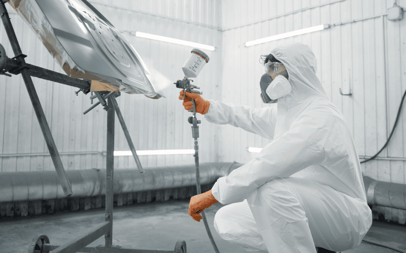 Person in a white protective suit and mask spray painting a car in a controlled environment.