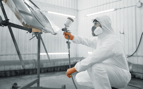 Person in a white protective suit and mask spray painting a car in a controlled environment.