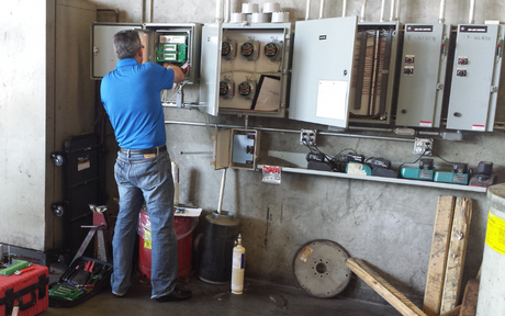 A worker in a blue shirt and jeans adjusts equipment inside open control panels on an industrial wall, surrounded by tools and machinery.