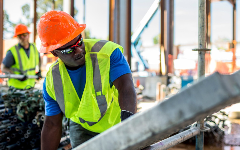 A construction worker wearing a bright yellow vest and orange hard hat focuses intently while operating equipment. Another worker is visible in the blurred background.