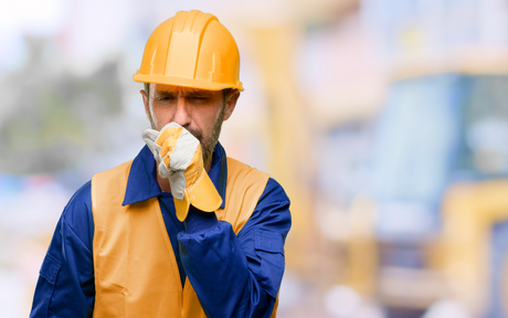 A construction worker in an orange hard hat and vest wipes his face with a gloved hand, appearing fatigued. Blurred machinery is visible in the background.