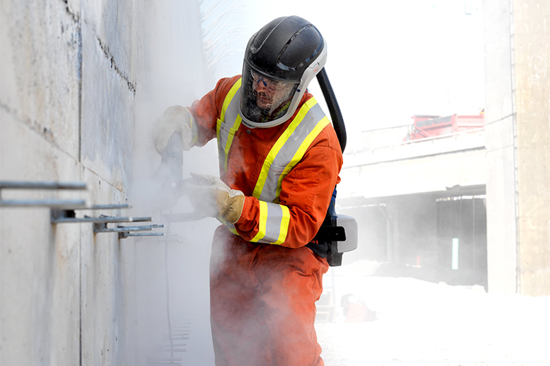 A worker in an orange safety suit and helmet uses a tool on a concrete wall, creating dust. The scene conveys diligence and industrial focus.