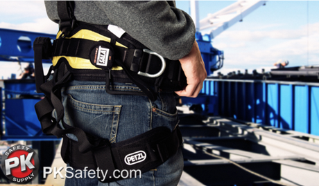 Closeup of worker wearing harness with Petzl logo near leg strap. Construction site in background.