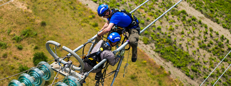 Two workers dressed in blue shirts with blue helmets, hang high on utility lines wearing harnesses.